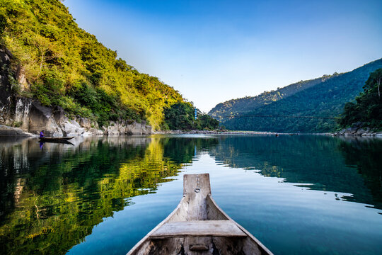 Tranquil wooden boat floating on crystal-clear turquoise waters of Umngot River in Dawki, Meghalaya with visible riverbed stones and lush green hills surrounding.