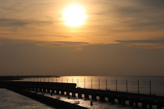 A majestic view of the Pamban Bridge during sunset, with its cantilever structure silhouetted against the vibrant orange and purple sky, reflecting on the calm waters of the Indian Ocean.