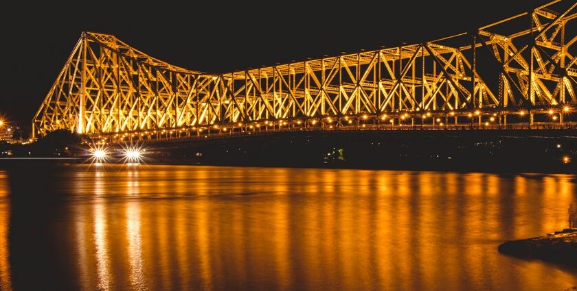 Howrah Bridge illuminated at night with city lights reflecting on the Hooghly River below.