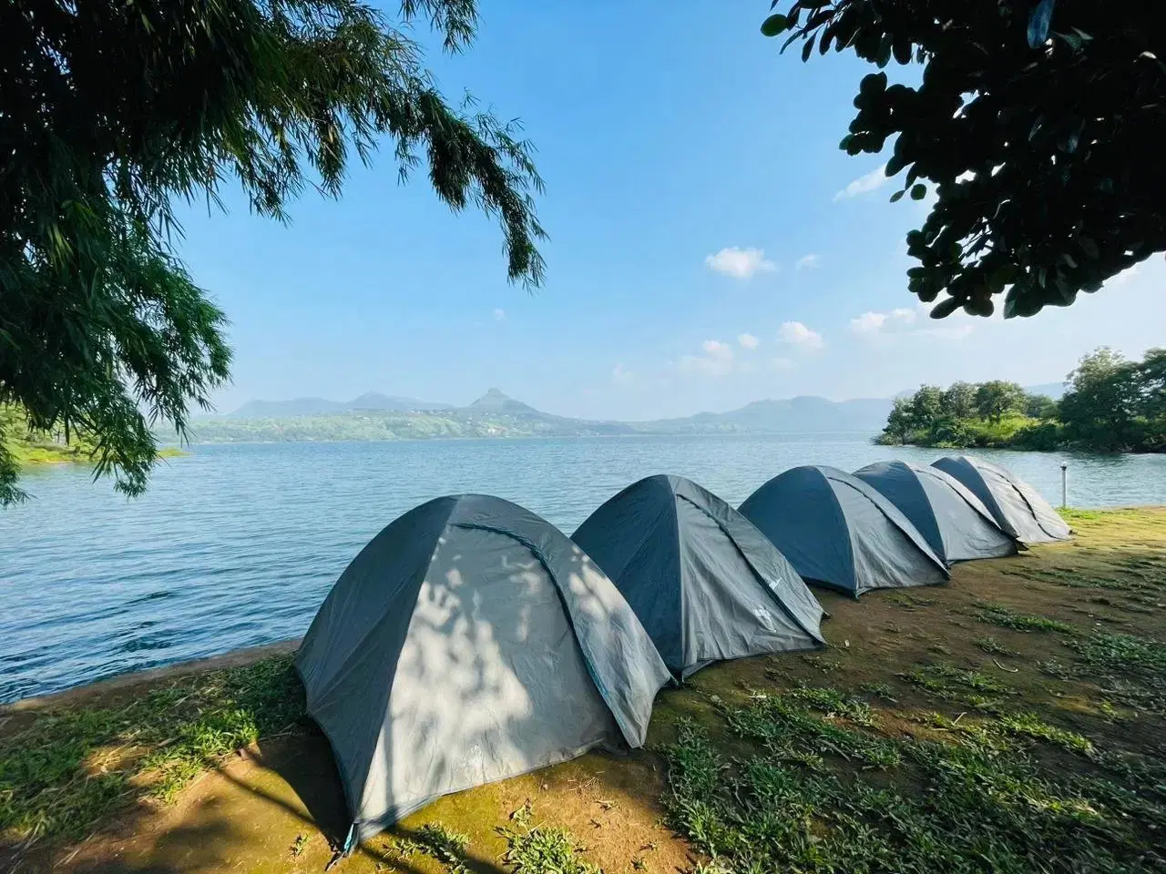A scenic campsite set up by the serene Pawna Lake, with tents glowing in the twilight and the Khandala hills in the distance.