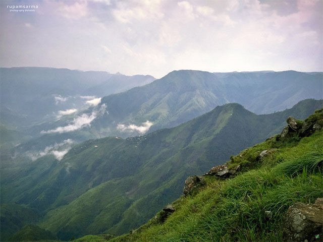 Panoramic view of steep grassy canyons with a winding river far below, mist rising from the valley, near Dawki
