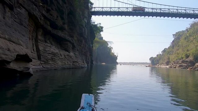 Tourist taking a photo of the iron bridge from the riverbank, with long shadows in late afternoon light, Dawki