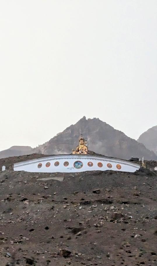 Distant golden Buddha statue perched atop a rocky hilltop plateau against a misty grey sky in Spiti Valley