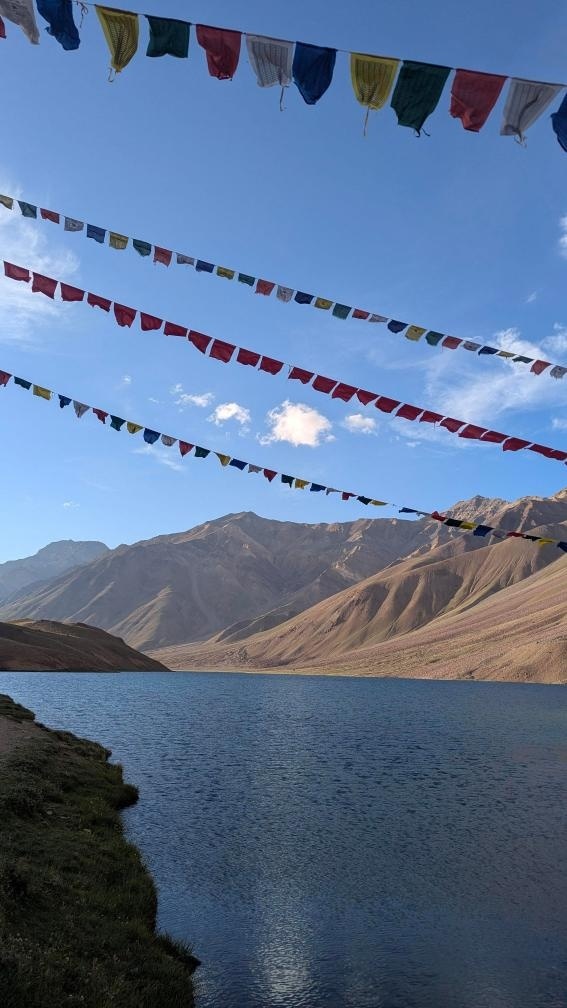 Multiple strings of colorful Buddhist prayer flags stretched across a vivid blue sky over the calm waters of Chandratal Lake
