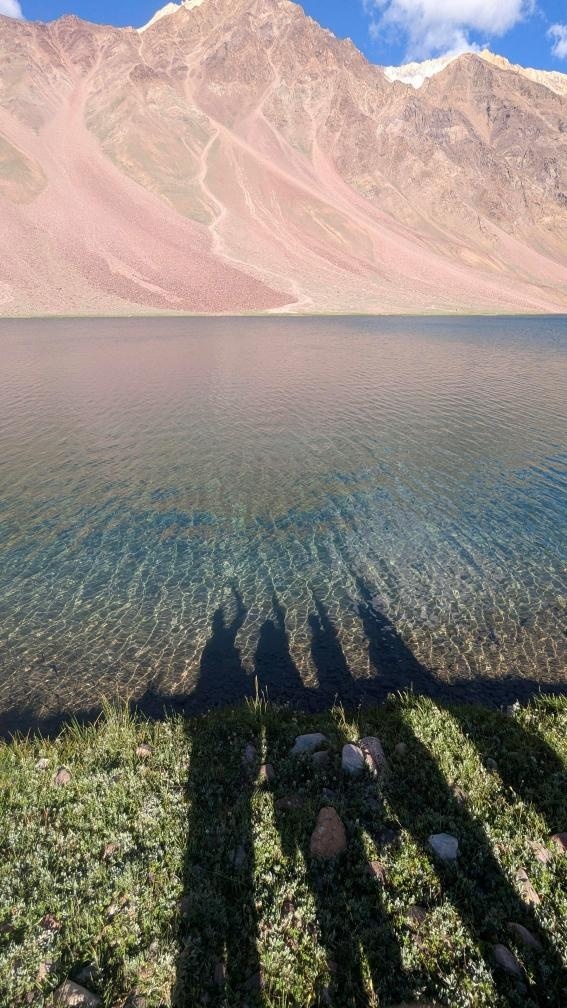 Shadows of travelers cast over the crystal clear shallow edge of Chandratal Lake with dramatic pink-hued barren mountains behind