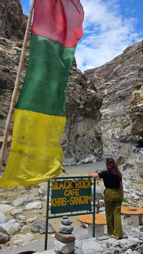 Traveler standing beneath colorful Buddhist prayer flags at Khab Sangam signboard where Spiti and Sutlej rivers meet