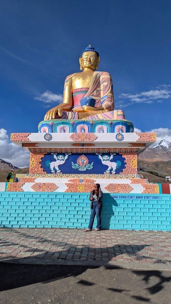 Traveler standing in front of the towering golden Langza Buddha statue with snow-capped mountains in the background, Spiti Valley
