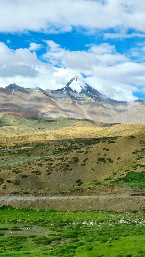 A sharp snow-capped Himalayan peak rising above golden barren hills and green meadows along the Spiti highway
